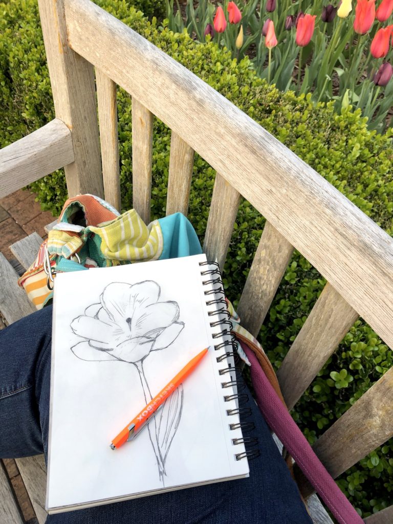 A sketchbook with a floral illustration and bag sitting on a wooden bench with tulips surrounding it.