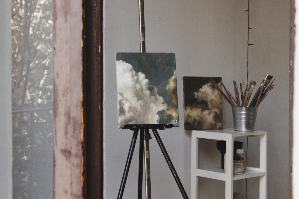 A black and white photo of a painting in progress. There is a standing easel with an abstract painting, and a small cart featuring various art supplies, which a tin can of paintbrushes on top.