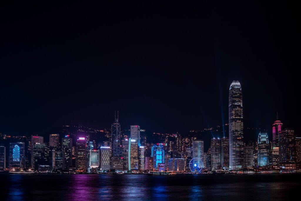 A city skyline lit up in cool blue and purple tones at night. Below the tall buildings is a body of water, and the lights from the buildings are reflected in the water.