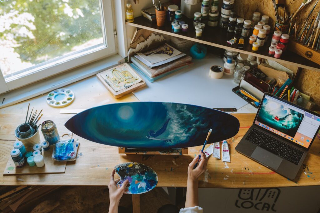 A painter working on a project with blue and green colors. There are notebooks and stacks of paint on a messy desk, and there is a laptop to the right of her with a reference image for her painting.