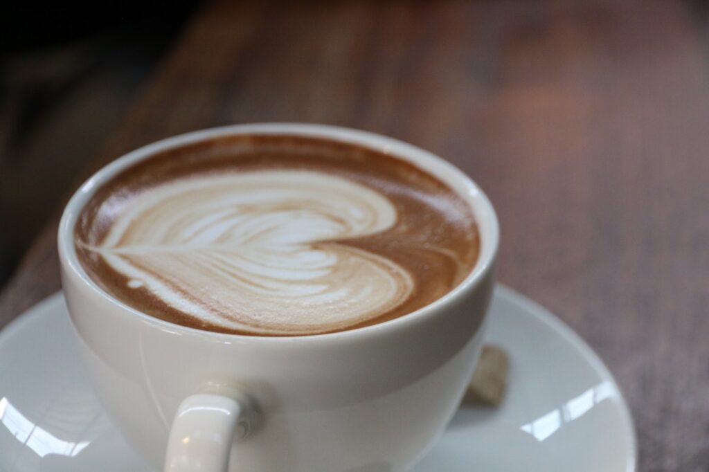 A white mug of coffee with latte art of a heart in the middle. The mug sits on top of a white plate, and is on a dark brown table.