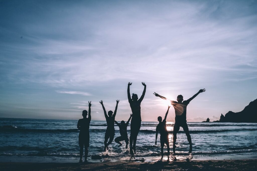 A silhouette of a group of friends hanging out at the beach. The ocean waves are just meeting their feet as the sun sets against a bright blue sky.