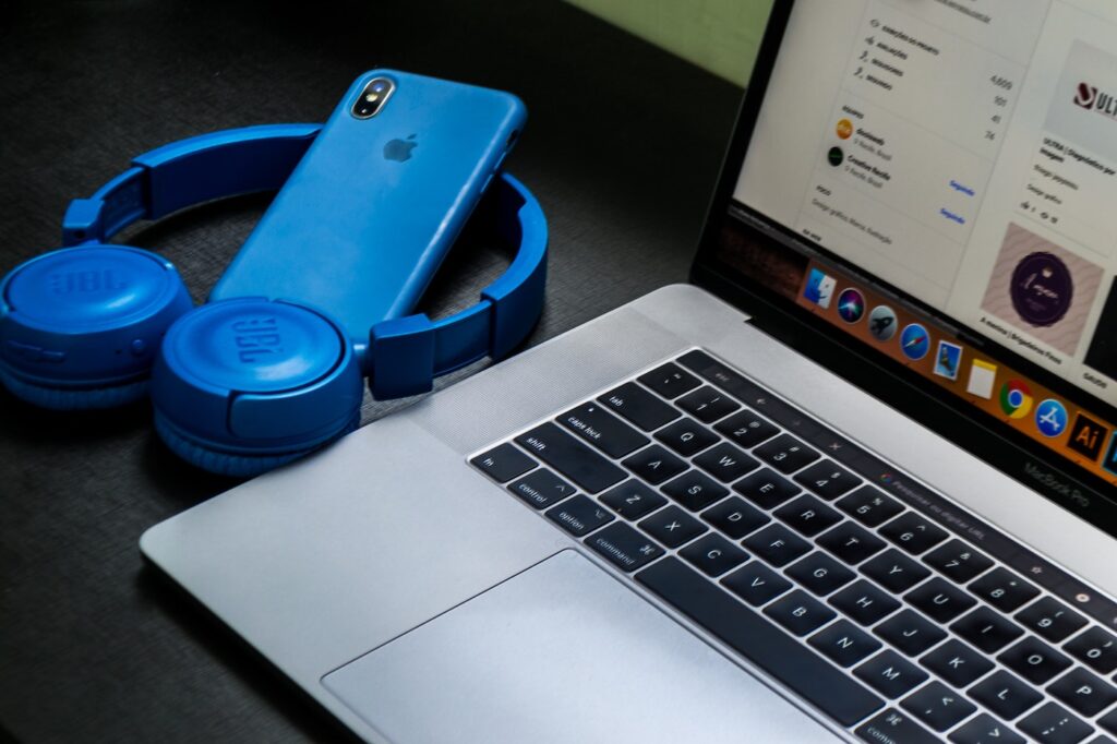 An open laptop with a blue iphone and pair of headphones sitting to the left. They are on top of a black table.