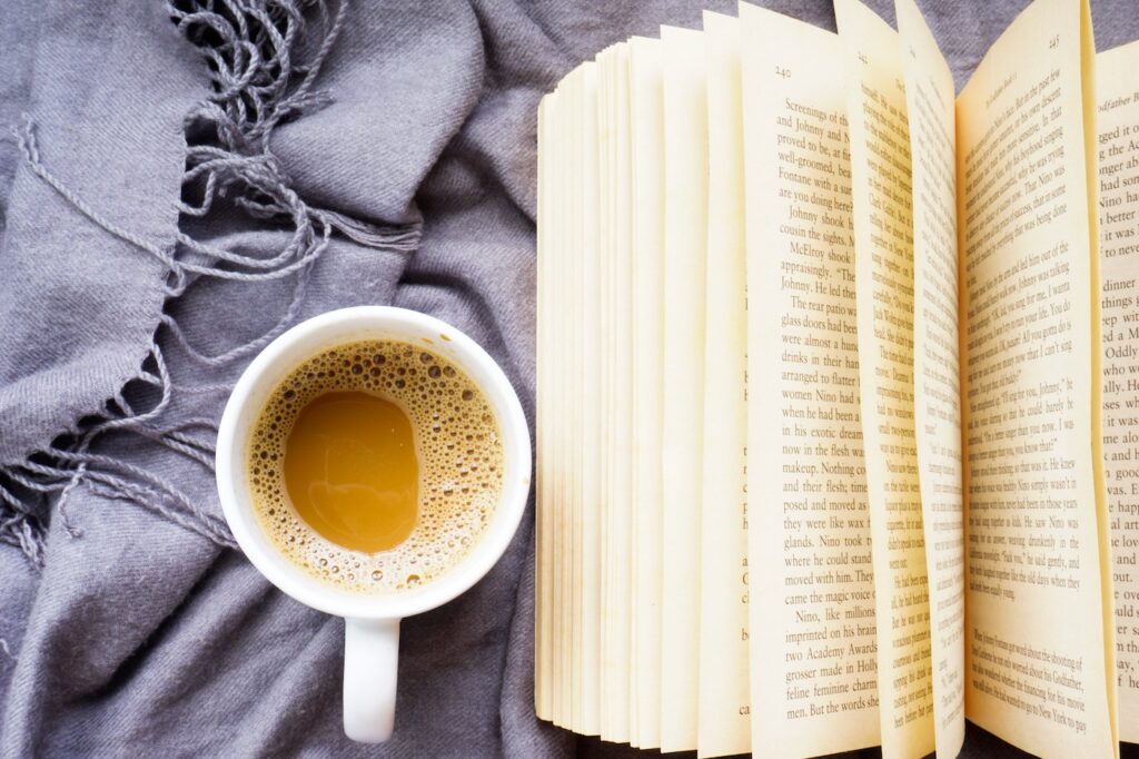 A white mug of warm tea, an open book, and a fuzzy beige blanket with fringe are laid out on a bed. There is also a whte sock peeking out of the blanket.