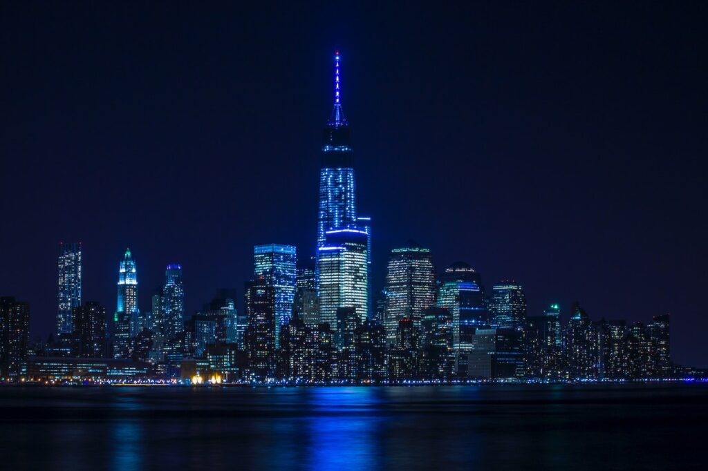 A city skyline lit with blue, pink, and purple lights sits against a night sky. The lights are reflected in a body of water below the buldings.