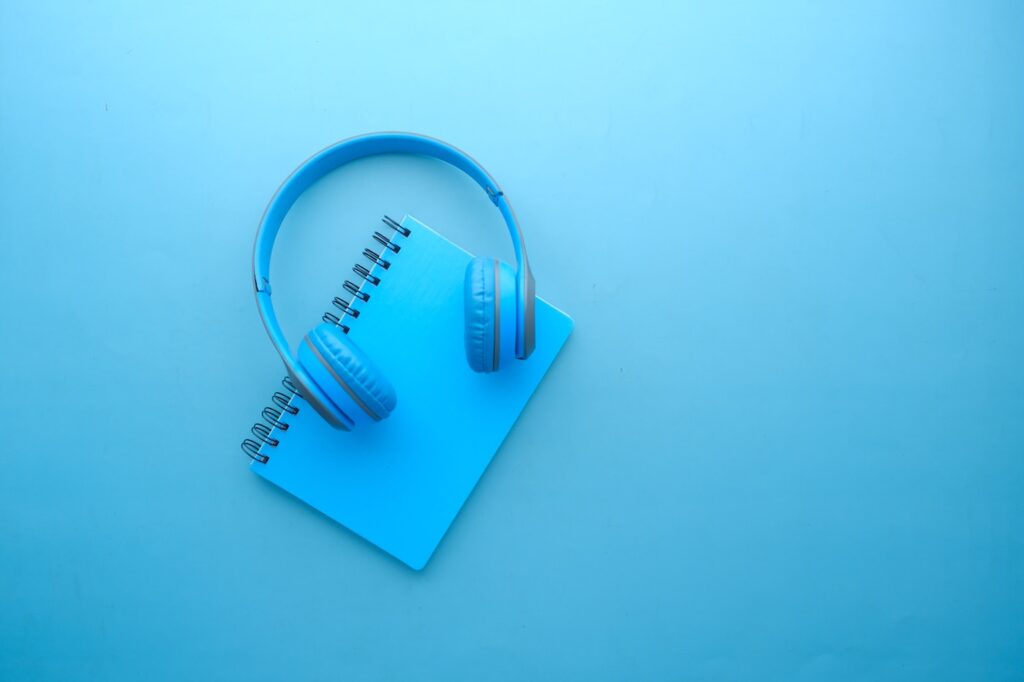 A notebook with a pair of headphones lying on top are set against a solid colored background. Everything in the photo is a sky blue.