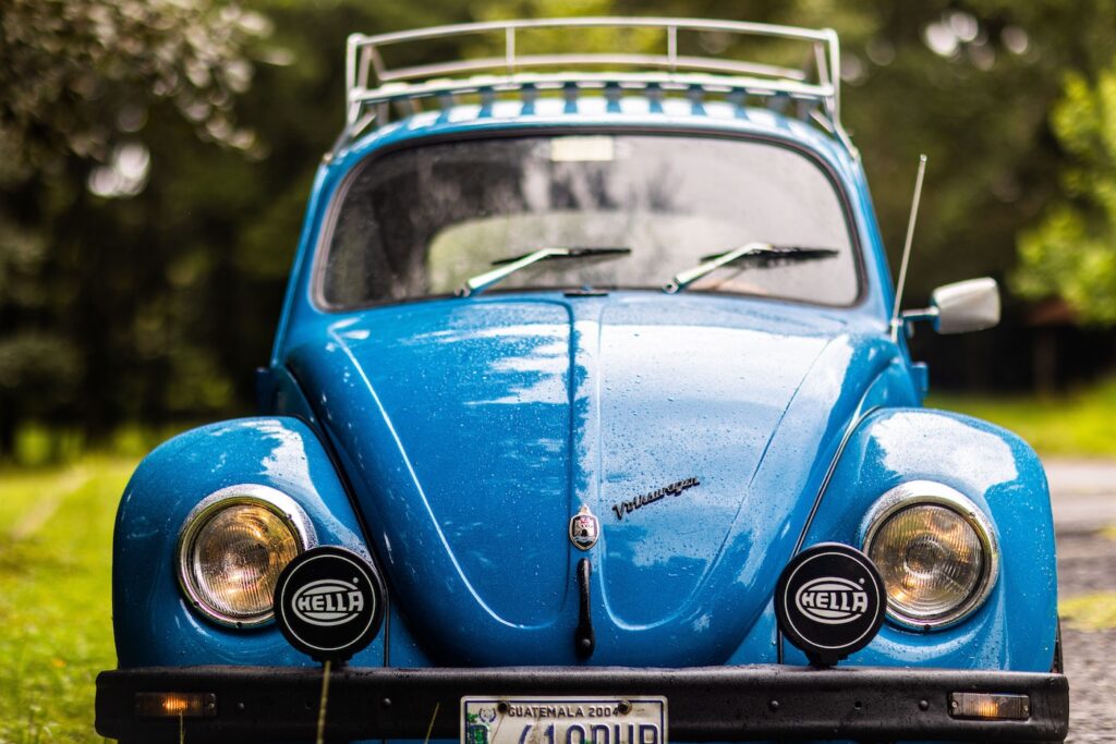 An old dark blue punch buggy convertible driving down a dirt road lined with green trees.