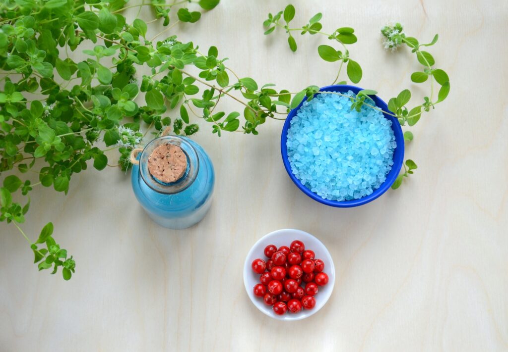 A blue bowl filled with blue colored salt and a white bowl of cherry tomatoes sit to the right of a bed of leafy herbs and a blue bottle with a cork. They are set on a white table.