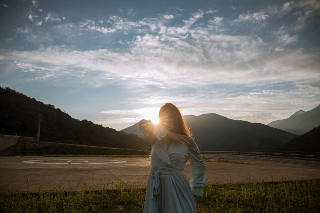 A young woman in a blue dress and dark hair has her face turned towards the sun. The sun is setting over a bunch of lush green hills, and is shining on the woman's head. The sky is a mix of pinks, purples, blues, and yellows.