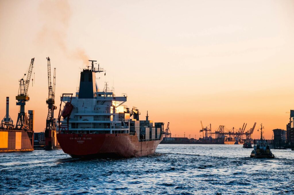 A large ship is setting out to sea, with an industrial shipyard in the background.