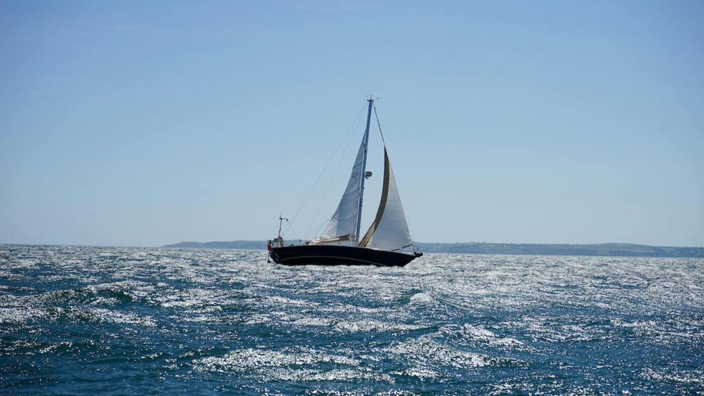 A single sailboat with a large white sail travels across a bright blue ocean with clear skies.