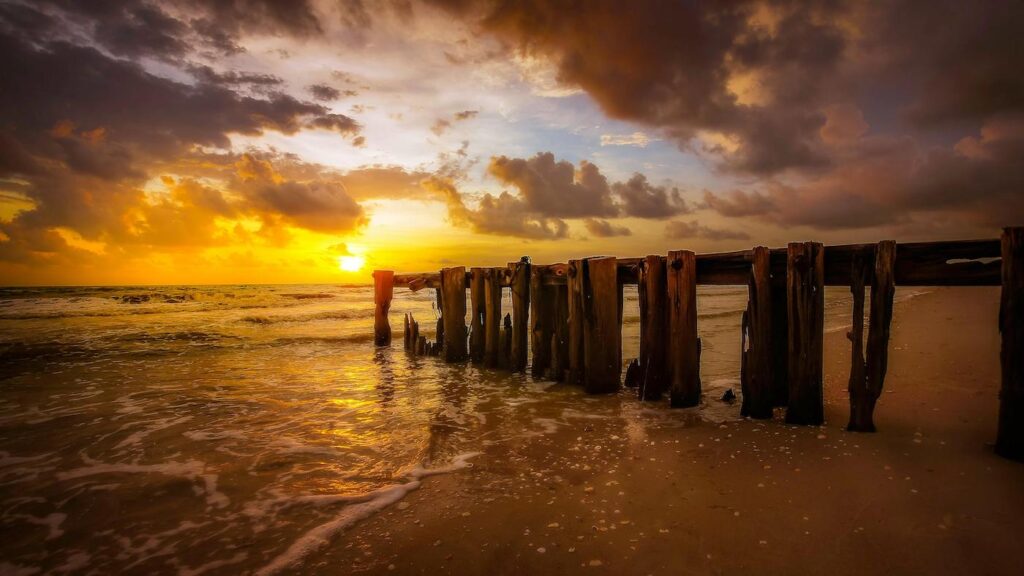 A long pier stretches across the ocean as the sun sets in the background.