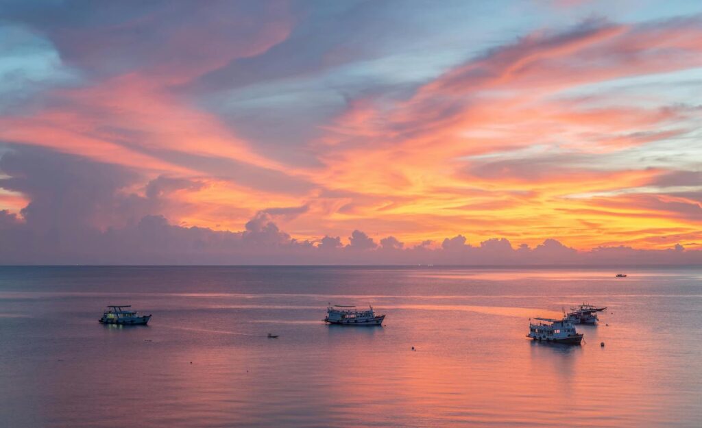 A group of small boats travel across the sea as the sun sets behind them.