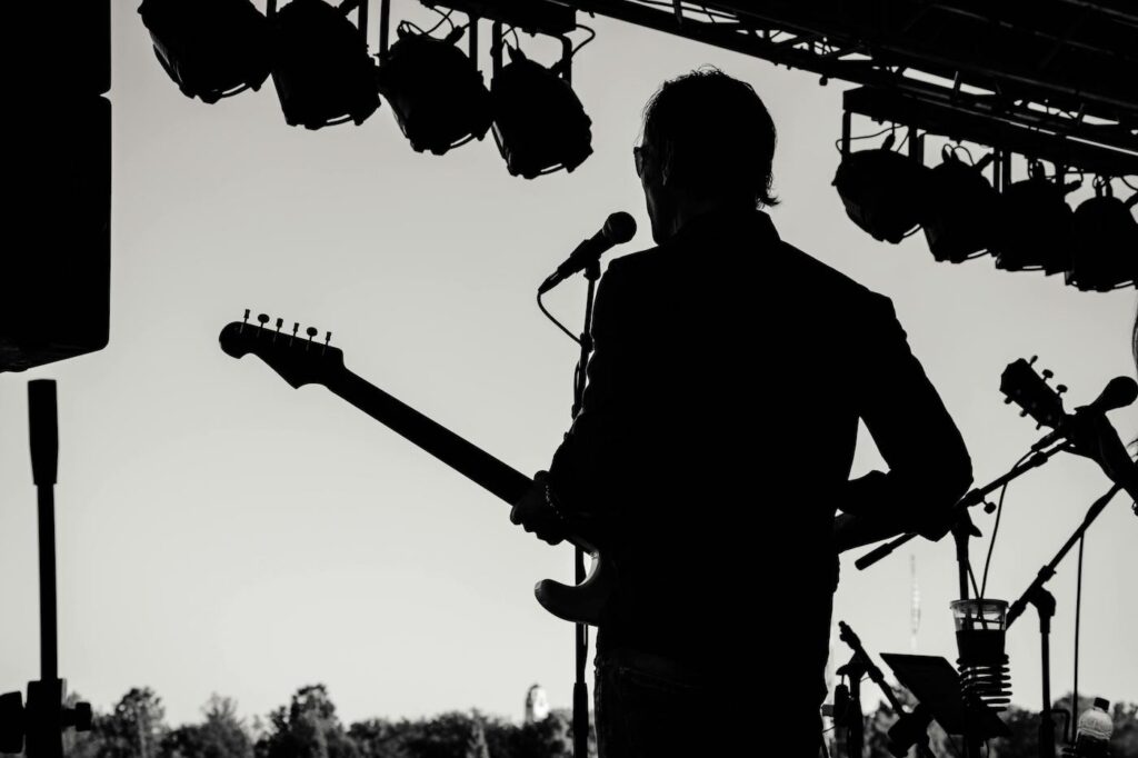 A man holding a guitar stands on stage in front of a microphone, performing outdoors at a concert venue.