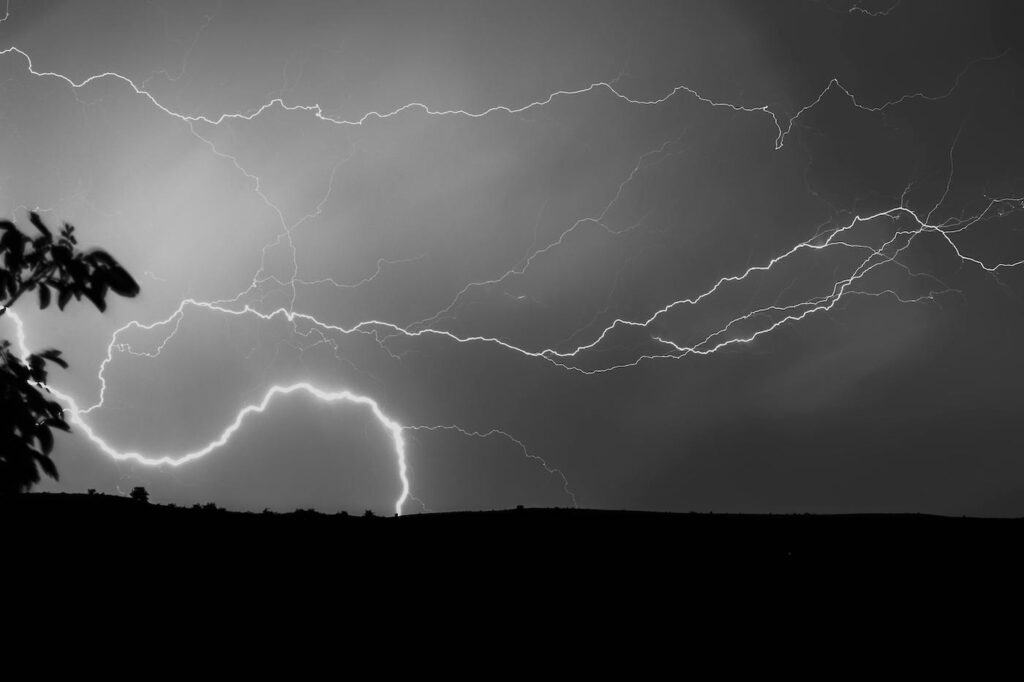 A dark and cloudy sky looms above  a field. Streaks of lightning fill the sky, with a bright bolt hitting the field.