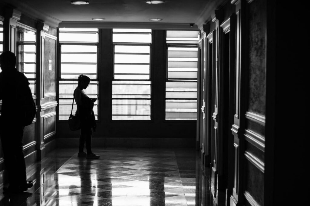 A woman looks down at her phone as she walks into a building's lobby. To the far left another person is walking towards the exit of the building.