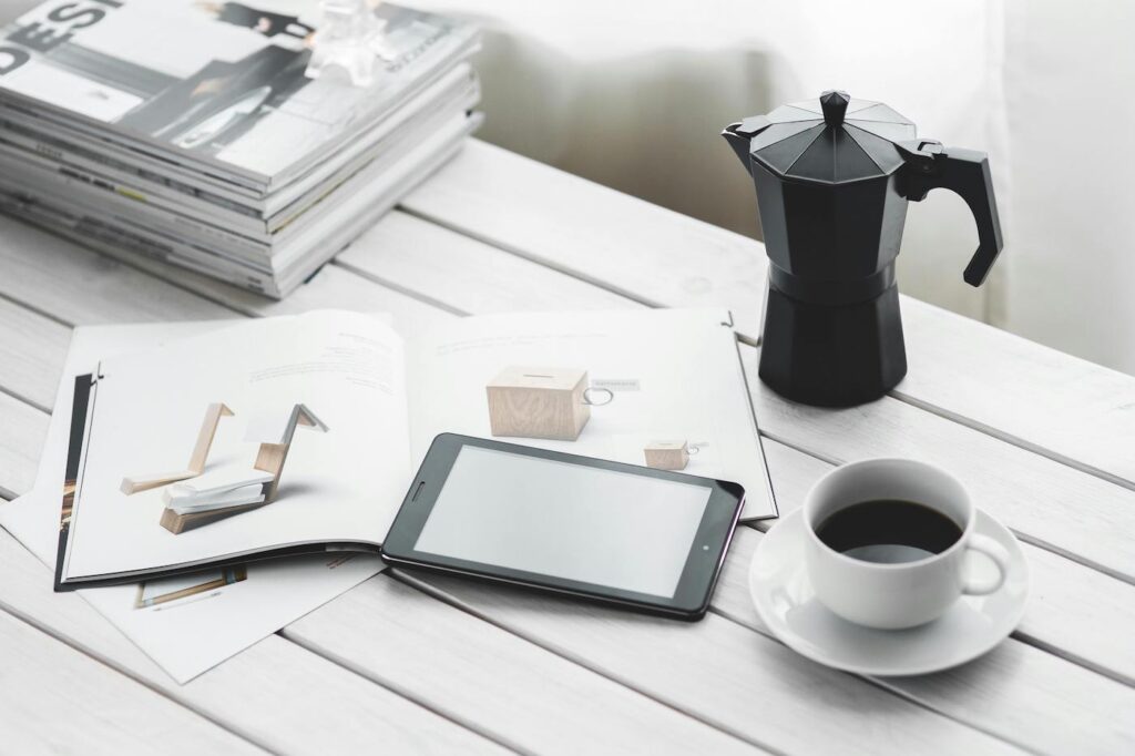 A wooden table with a stack of books to the left, and a pot of coffee to the right. Near the bottom center is an open book, an iphone, and a white mug full of coffee.