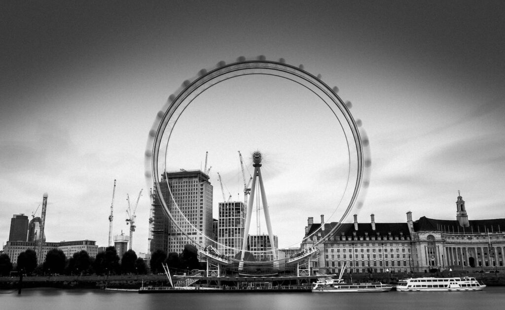 A wide shot of the London Eye and the surrounding environment, which includes a stretch of buildings and a long river with ferry boats docked nearby.